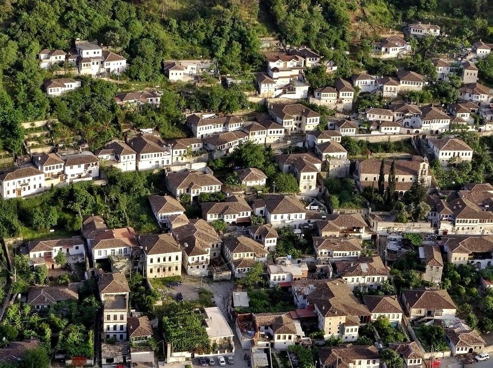 An aerial view of a densely packed residential area with white houses and red-tiled roofs, surrounded by greenery.