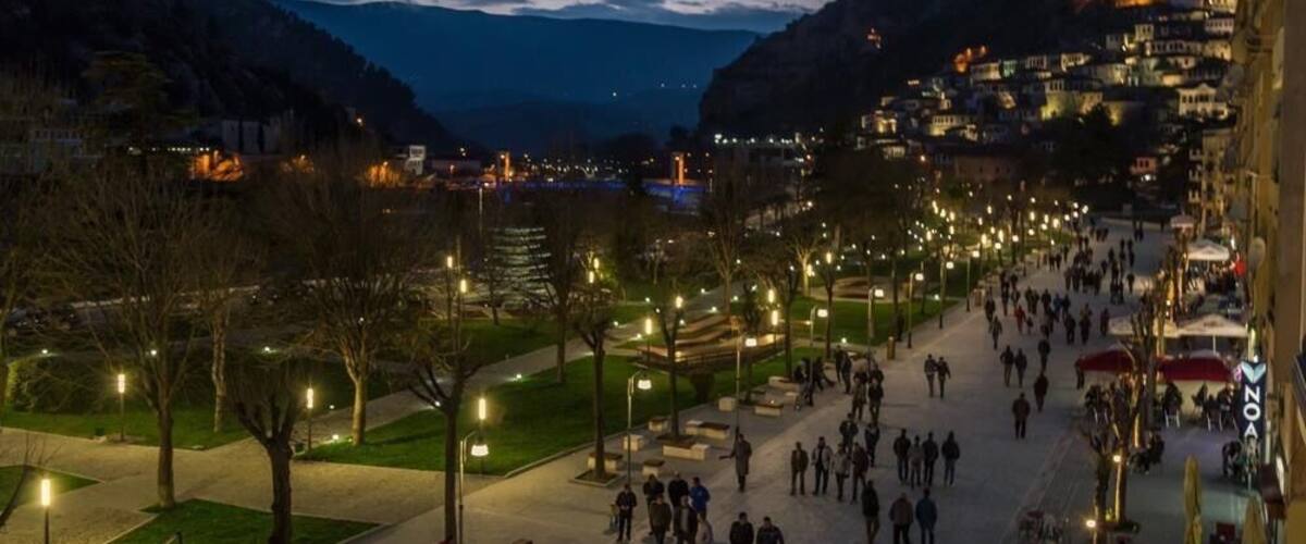A nighttime cityscape with a river, illuminated buildings, and a park with people.