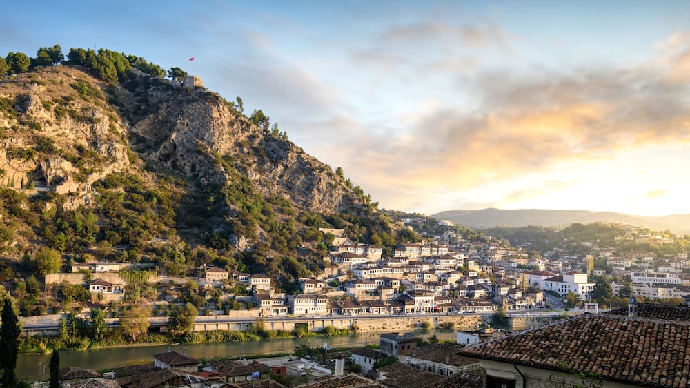 A town nestled between hills with a river, historic buildings, and a flag on a hilltop.
