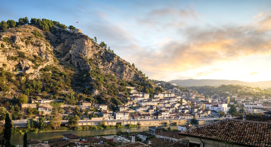 Un pueblo situado entre colinas con un río, edificios históricos y una bandera en la cima.