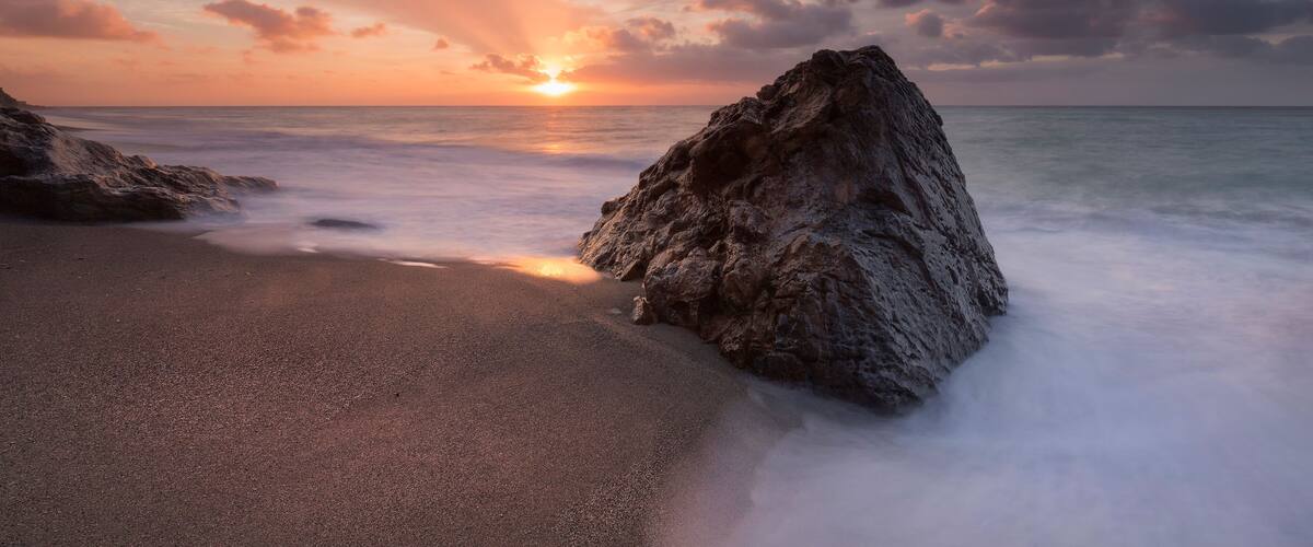 A sunset over a rocky coastline with waves gently lapping at the shore.