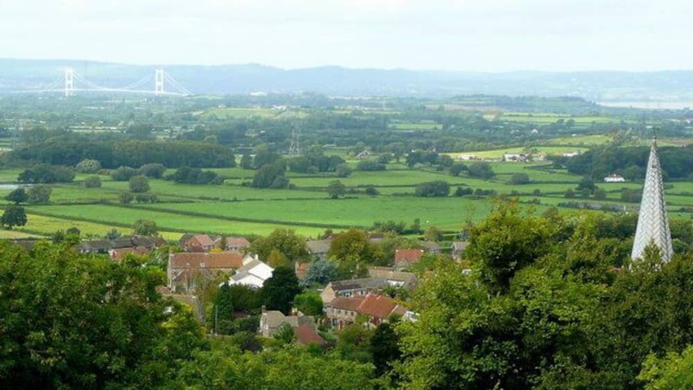 Un paysage rural avec le clocher d’une église, des champs verdoyants ondulants et un pont situé au loin.