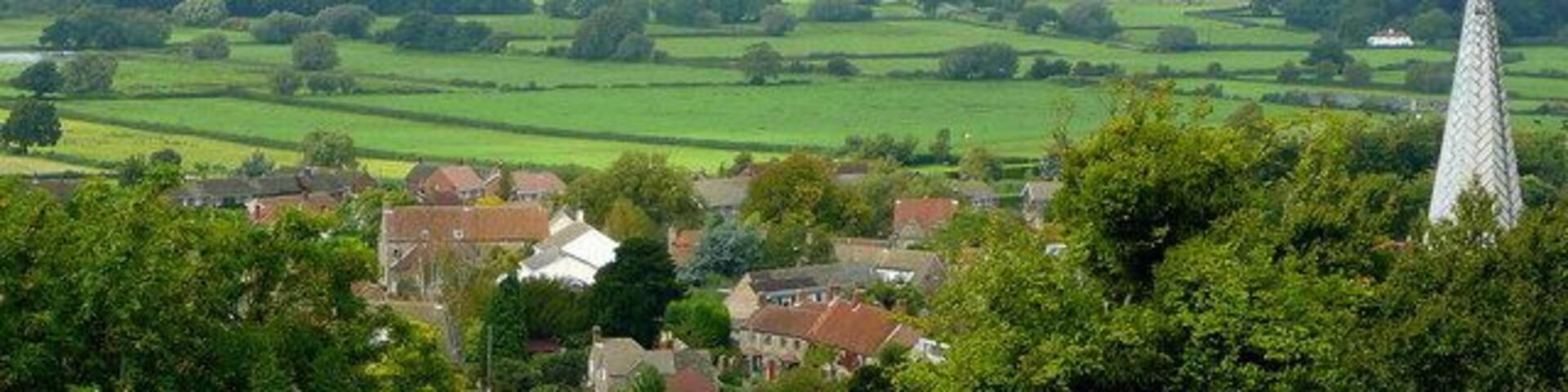 A rural landscape with a church spire, rolling green fields, and a distant bridge.