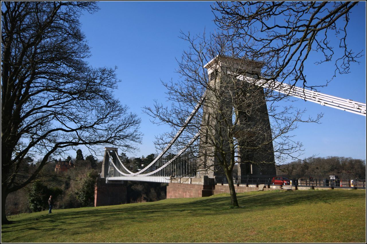 A suspension bridge with a tower, surrounded by trees and a grassy area.