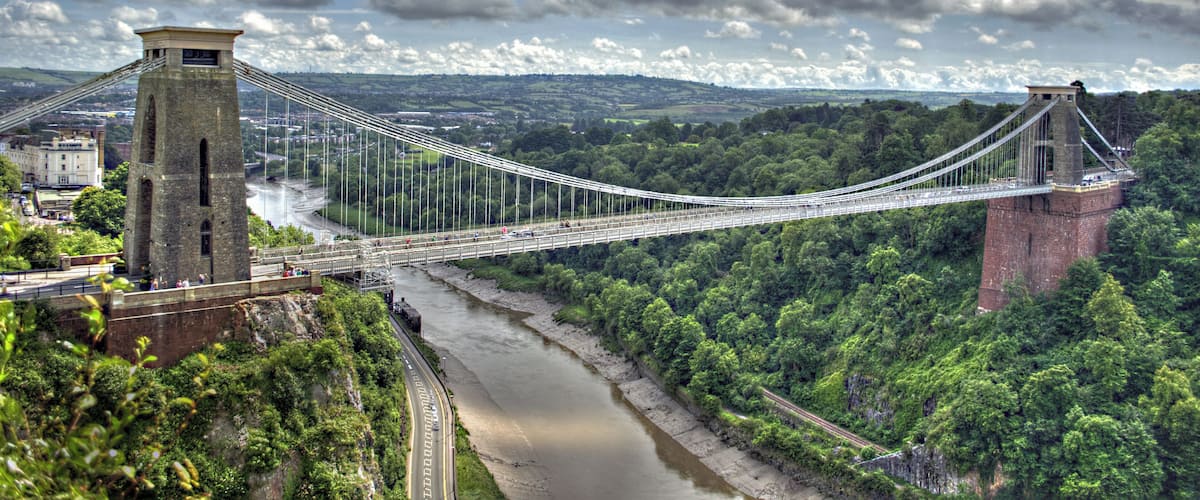 The Clifton Suspension Bridge spans the Avon Gorge and links Clifton in Bristol to Leigh Woods in North Somerset, England. Designed by Isambard Kingdom Brunel, the bridge is a grade I listed building. The idea of building a bridge across the Avon Gorge originated in 1753. The original plans were for a stone bridge, and later plans were for a cast iron structure. An attempt to build Brunel's design in 1831 was stopped by the Bristol Riots, and the revised version of his designs was built after his death, and completed in 1864. The bridge is a distinctive landmark, used as a symbol of Bristol on postcards, promotional materials, and informational web sites. It has also been used as a backdrop to several films and television programmes.