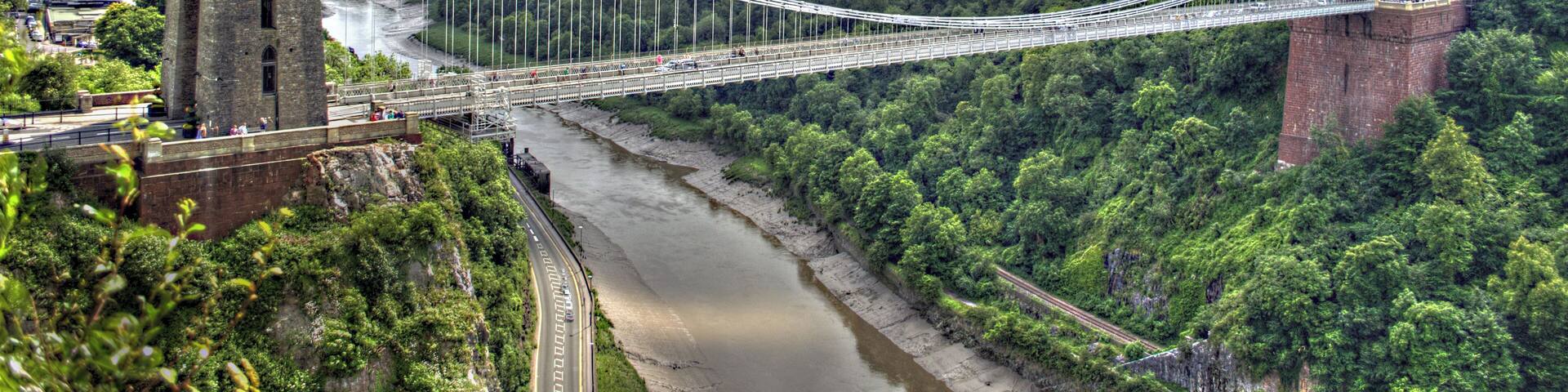 The Clifton Suspension Bridge spans the Avon Gorge and links Clifton in Bristol to Leigh Woods in North Somerset, England. Designed by Isambard Kingdom Brunel, the bridge is a grade I listed building. The idea of building a bridge across the Avon Gorge originated in 1753. The original plans were for a stone bridge, and later plans were for a cast iron structure. An attempt to build Brunel's design in 1831 was stopped by the Bristol Riots, and the revised version of his designs was built after his death, and completed in 1864. The bridge is a distinctive landmark, used as a symbol of Bristol on postcards, promotional materials, and informational web sites. It has also been used as a backdrop to several films and television programmes.