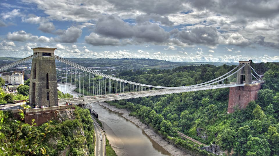 The Clifton Suspension Bridge spans the Avon Gorge and links Clifton in Bristol to Leigh Woods in North Somerset, England. Designed by Isambard Kingdom Brunel, the bridge is a grade I listed building. The idea of building a bridge across the Avon Gorge originated in 1753. The original plans were for a stone bridge, and later plans were for a cast iron structure. An attempt to build Brunel's design in 1831 was stopped by the Bristol Riots, and the revised version of his designs was built after his death, and completed in 1864. The bridge is a distinctive landmark, used as a symbol of Bristol on postcards, promotional materials, and informational web sites. It has also been used as a backdrop to several films and television programmes.