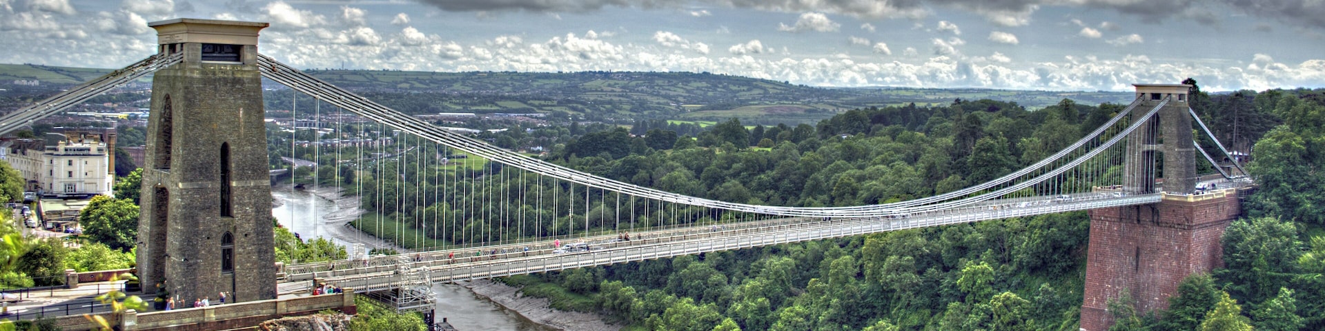 The Clifton Suspension Bridge spans the Avon Gorge and links Clifton in Bristol to Leigh Woods in North Somerset, England. Designed by Isambard Kingdom Brunel, the bridge is a grade I listed building. The idea of building a bridge across the Avon Gorge originated in 1753. The original plans were for a stone bridge, and later plans were for a cast iron structure. An attempt to build Brunel's design in 1831 was stopped by the Bristol Riots, and the revised version of his designs was built after his death, and completed in 1864. The bridge is a distinctive landmark, used as a symbol of Bristol on postcards, promotional materials, and informational web sites. It has also been used as a backdrop to several films and television programmes.