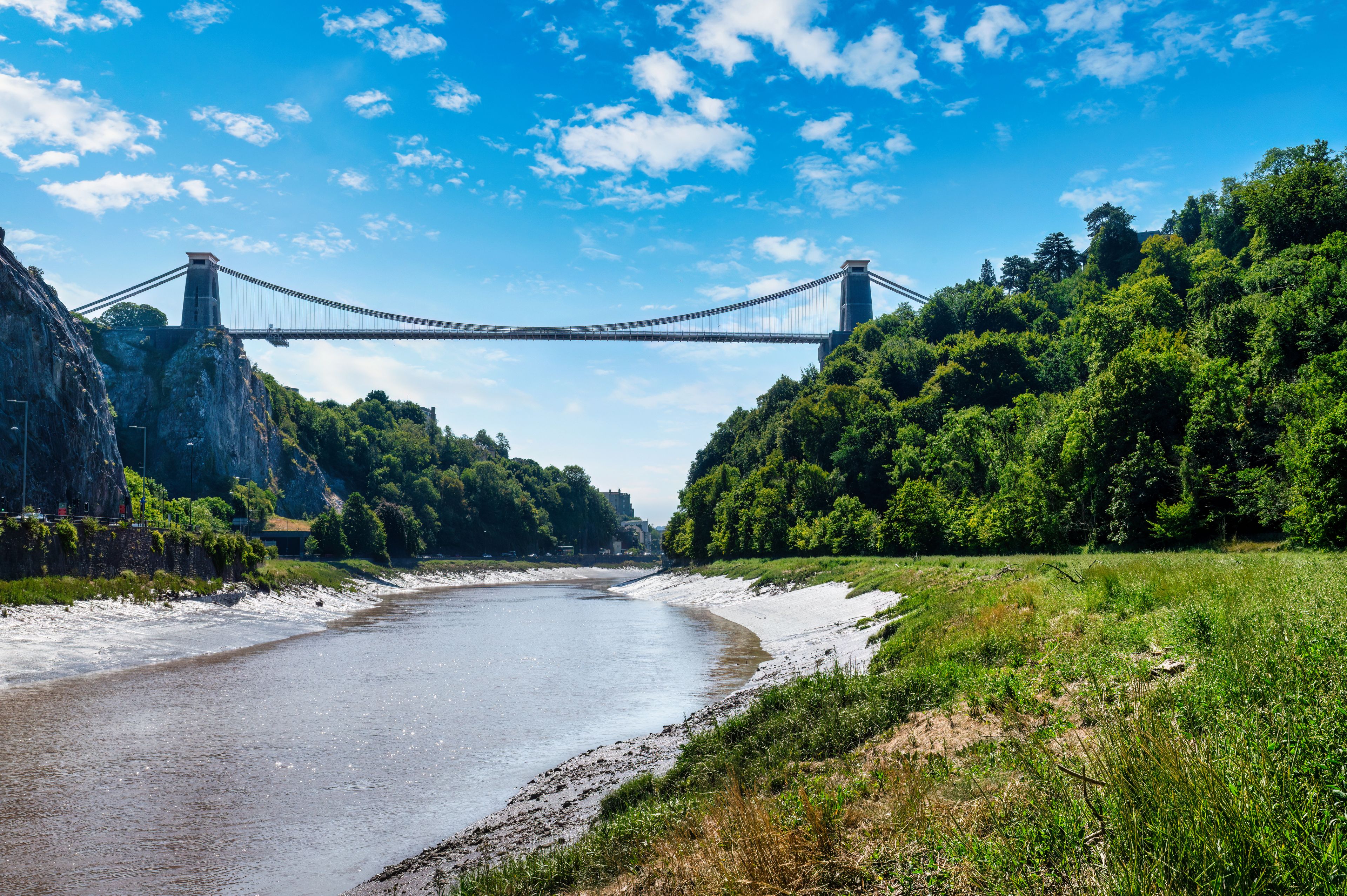 Une rivière traversée par un pont suspendu, bordée d’une végétation luxuriante et de falaises rocheuses.