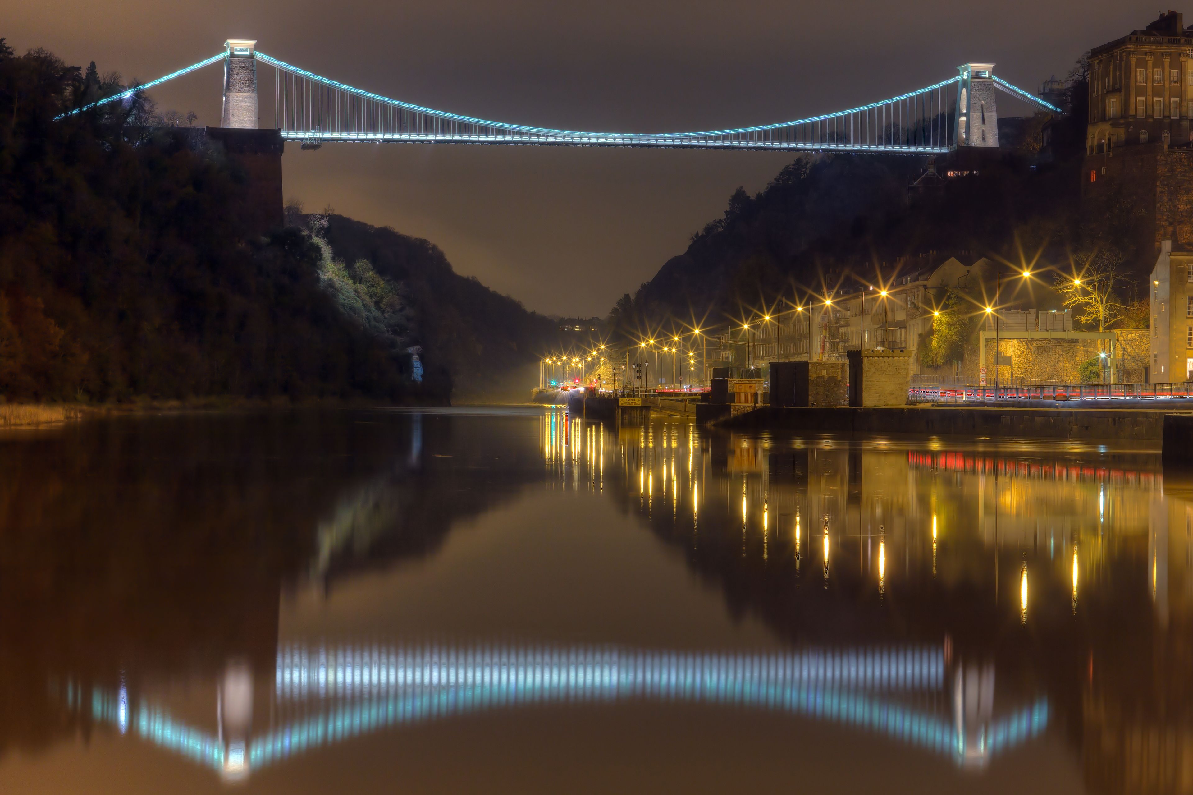 Un pont illuminé dont la lumière se reflète sur une rivière paisible, entouré d’arbres et de bâtiments.
