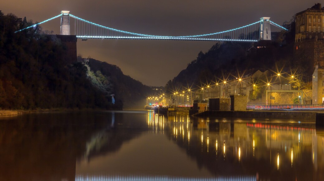Un pont illuminé dont la lumière se reflète sur une rivière paisible, entouré d’arbres et de bâtiments.