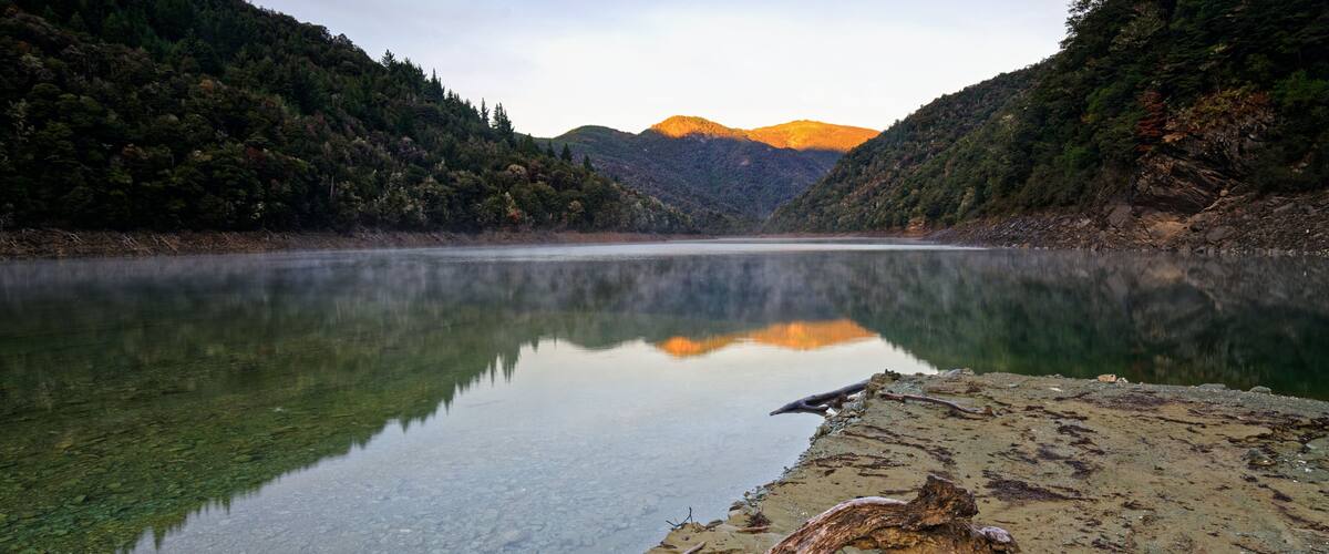 A calm lake with clear water, surrounded by forested hills and a mountain in the background.