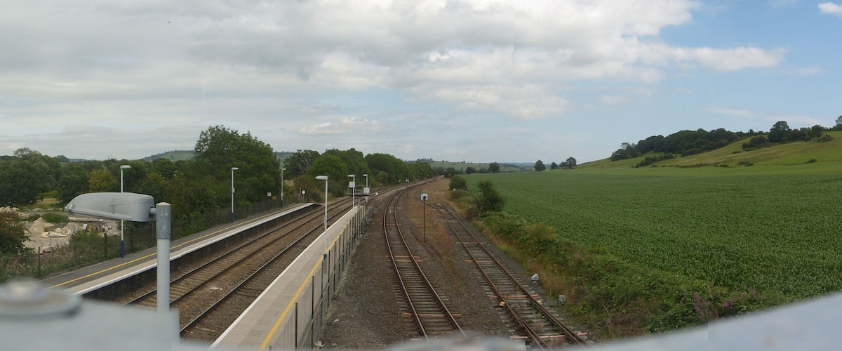 A railway track with a platform, surrounded by green fields and trees under a cloudy sky.