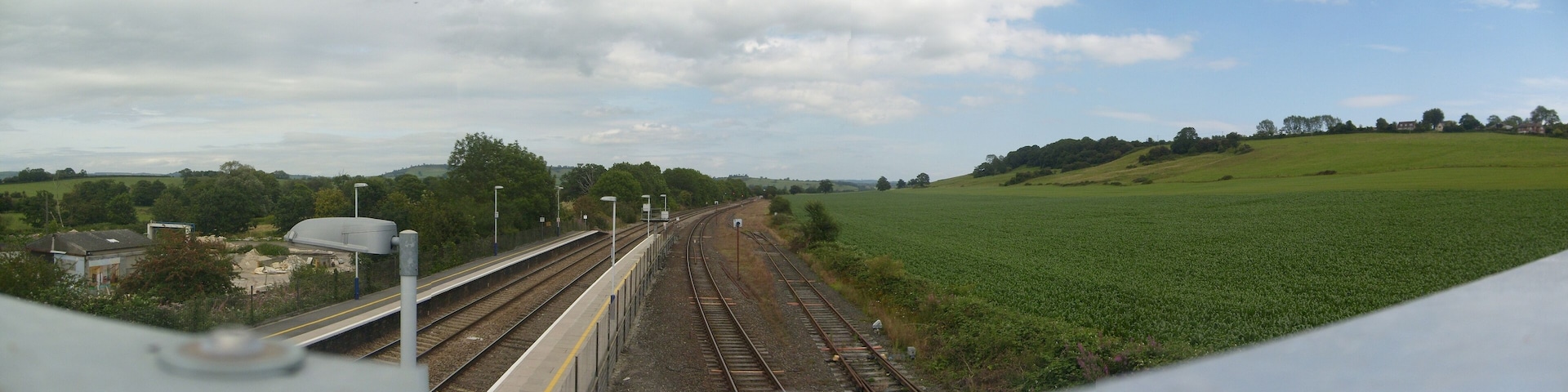 A railway track with a platform, surrounded by green fields and trees under a cloudy sky.