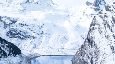 A person in a red jacket standing by a calm lake surrounded by snow-covered mountains.
