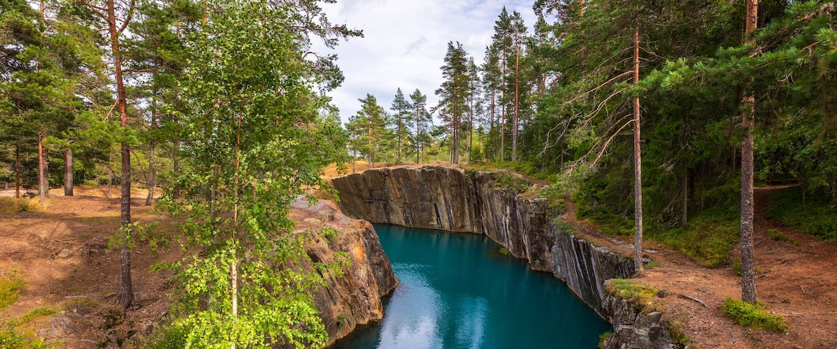 Aerial view of the flooded mine pit at Ostra Silvberg gruva with turquoise water surrounded by pine forest and rocky cliffs in Borlange, Dalarna County, Sweden.