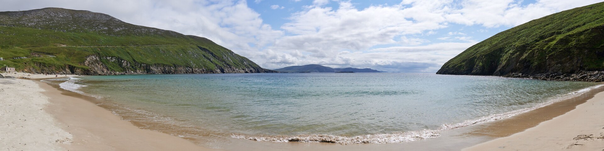 A beach with clear turquoise waters, green hills, and a distant mountain range.