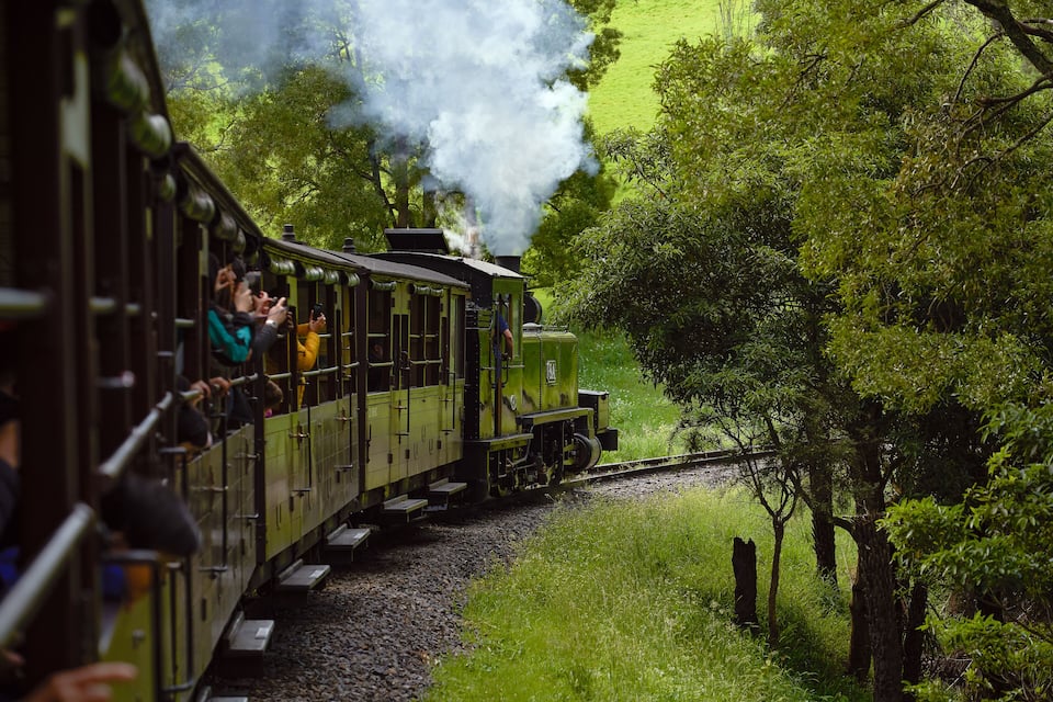 A green steam train with passengers, traveling through a lush green landscape with trees and a river.