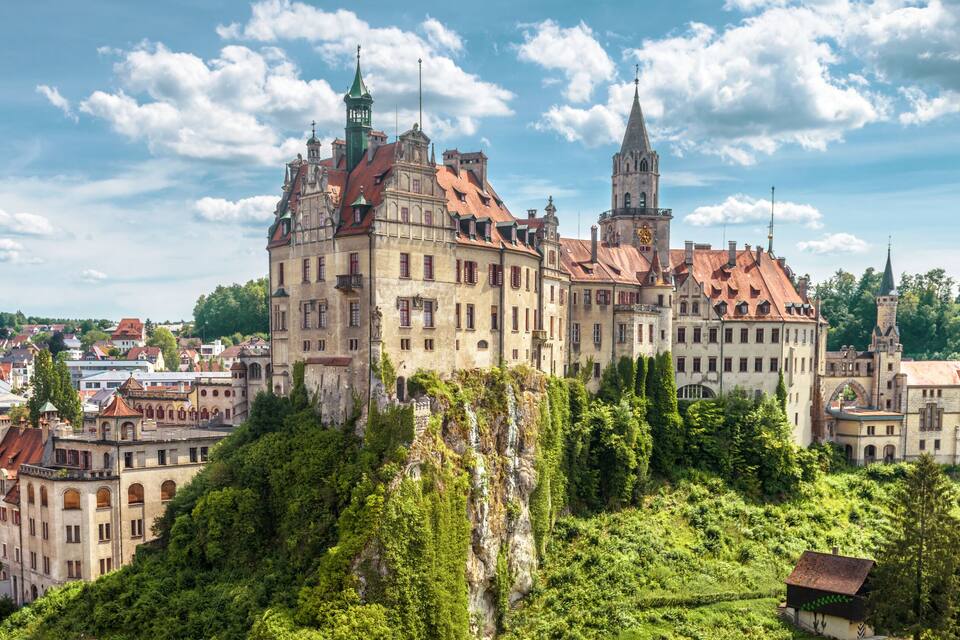 A historic castle with multiple towers, surrounded by lush greenery and a town below.