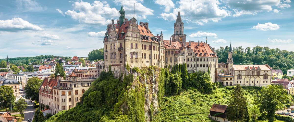 A historic castle with multiple towers, surrounded by lush greenery and a town below.