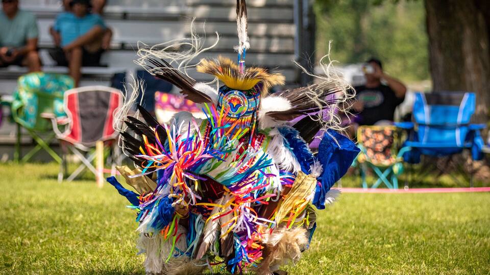 A person in a colorful traditional costume with feathers and ribbons, standing on grass with spectators in the background.
