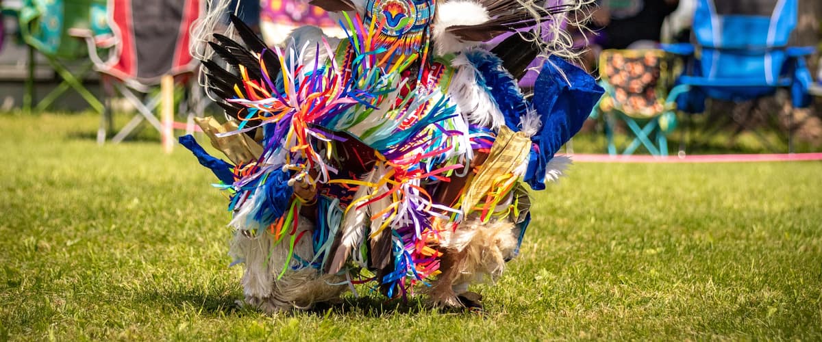 A person in a colorful traditional costume with feathers and ribbons, standing on grass with spectators in the background.
