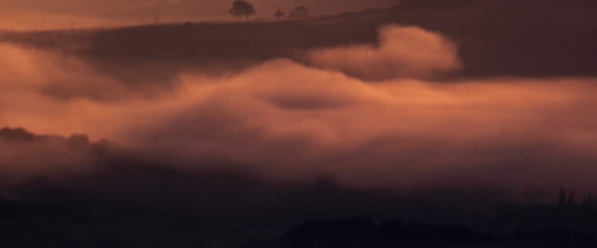 A landscape with rolling hills and fields covered in mist during sunrise or sunset.