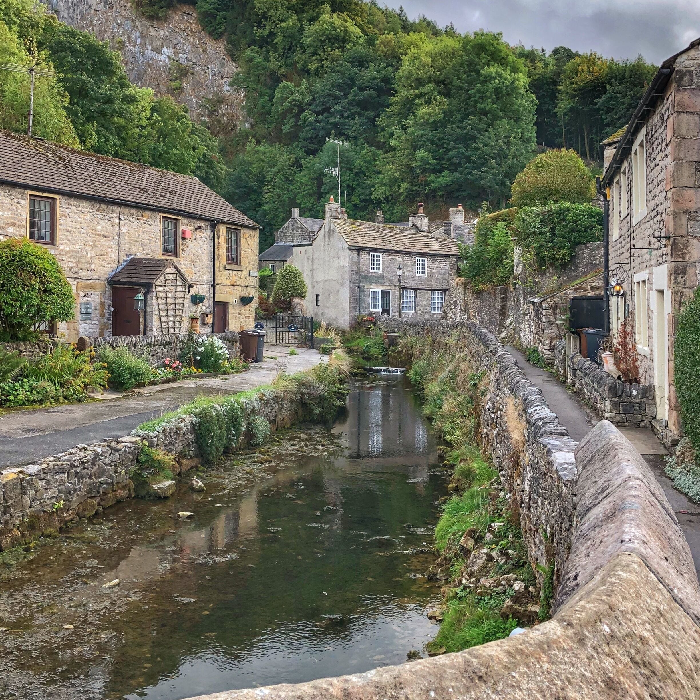 A narrow waterway flanked by stone buildings, a stone wall, and lush greenery.