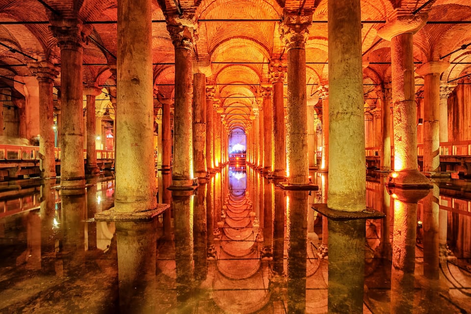 An interior with a reflective floor, large columns, and a central illuminated structure.