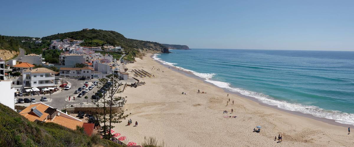 A coastal town with a sandy beach, a clear blue sea, and buildings on a hillside.