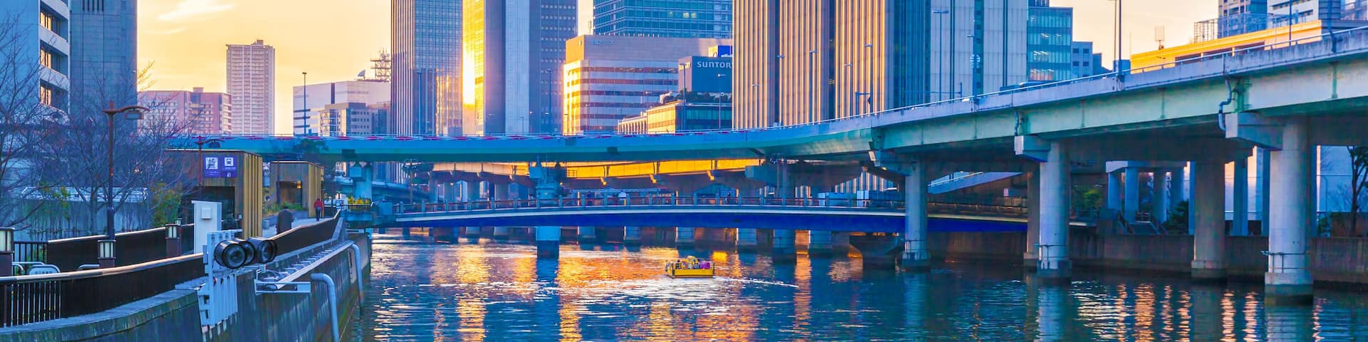 A city skyline with modern skyscrapers, a river reflecting the sunset, and a bridge crossing over the water.