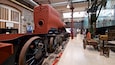 A large red steam engine in a museum with historical displays and a warning sign.