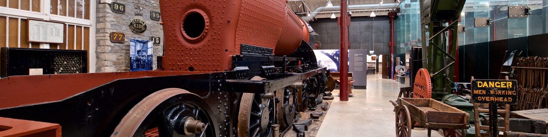 A large red steam engine in a museum with historical displays and a warning sign.