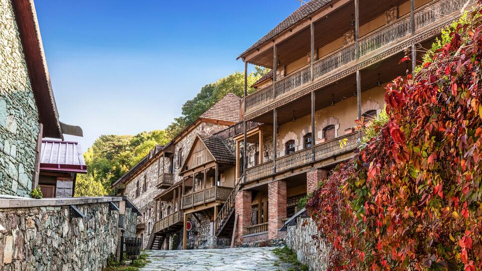 A cobblestone street flanked by traditional wooden buildings with balconies, a stone wall, and a backdrop of trees.