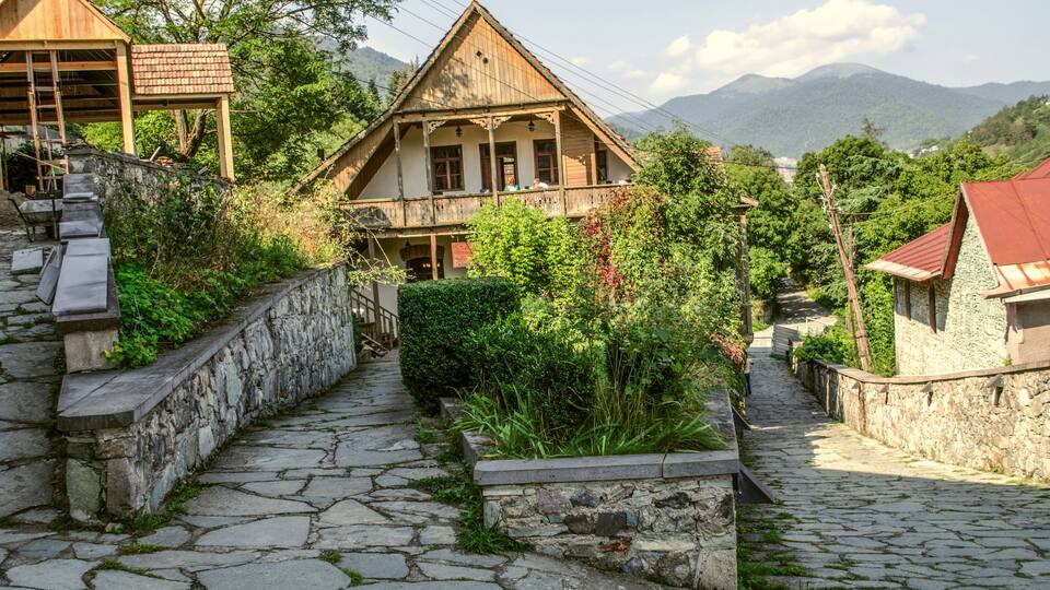 A stone-paved village street with traditional wooden houses, a stone wall, and a mountain backdrop.