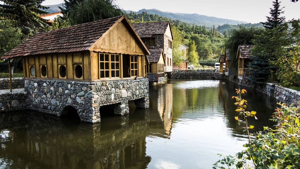 Wooden houses with stone bases by a calm river, surrounded by lush greenery and mountains.