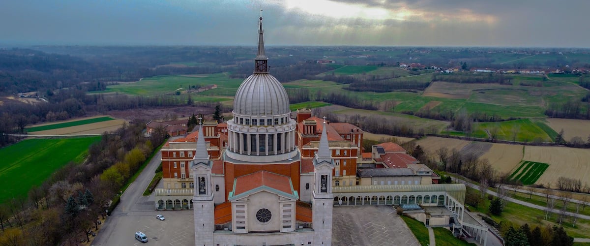 An aerial view of a large church with a prominent dome and bell tower, surrounded by green fields and a few scattered buildings.