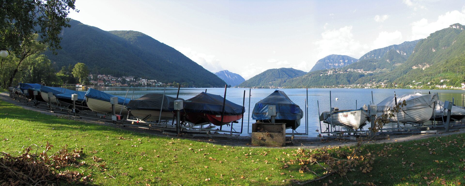 A lakeside with boats covered in tarps, mountains in the background, and a grassy area with fallen leaves.