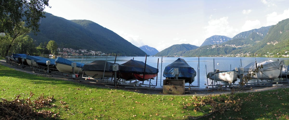 A lakeside with boats covered in tarps, mountains in the background, and a grassy area with fallen leaves.