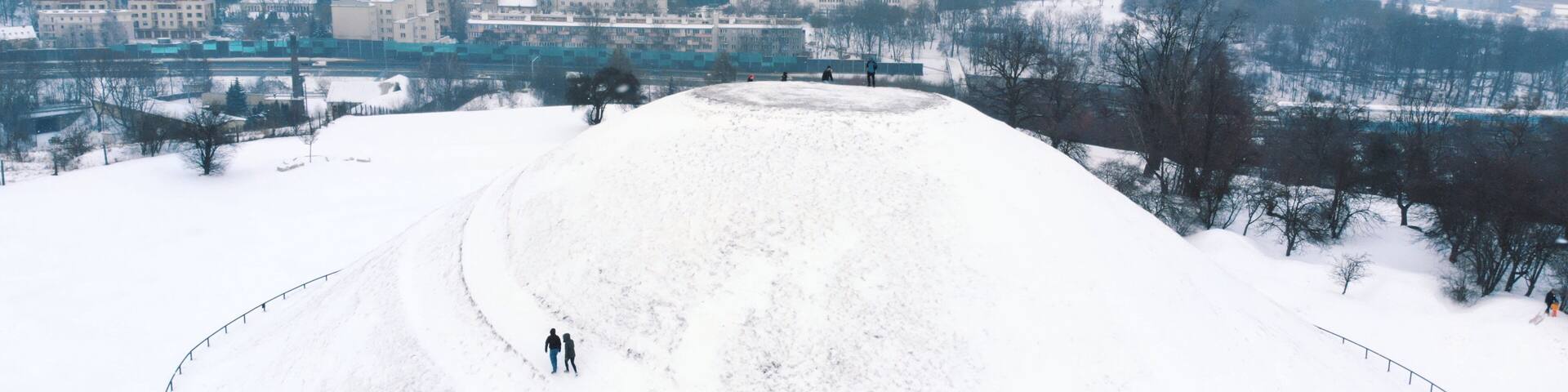 A snow-covered hill with a few people walking. In the background, there are buildings and trees.