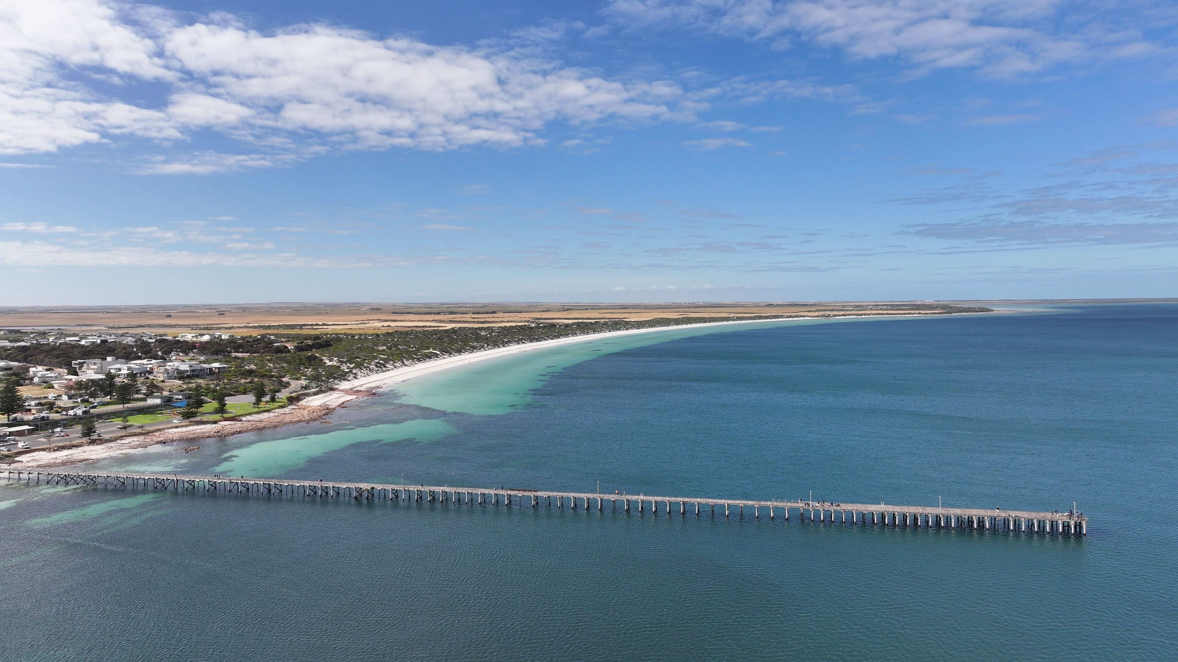 A coastal town with a long pier extending into the ocean, a sandy beach, and a clear blue sky.