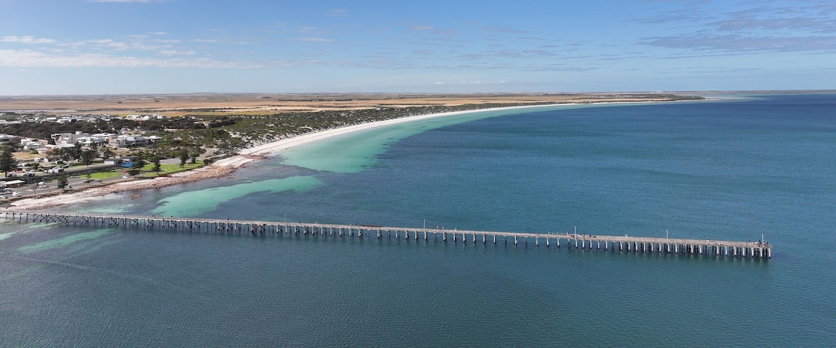 A coastal town with a long pier extending into the ocean, a sandy beach, and a clear blue sky.