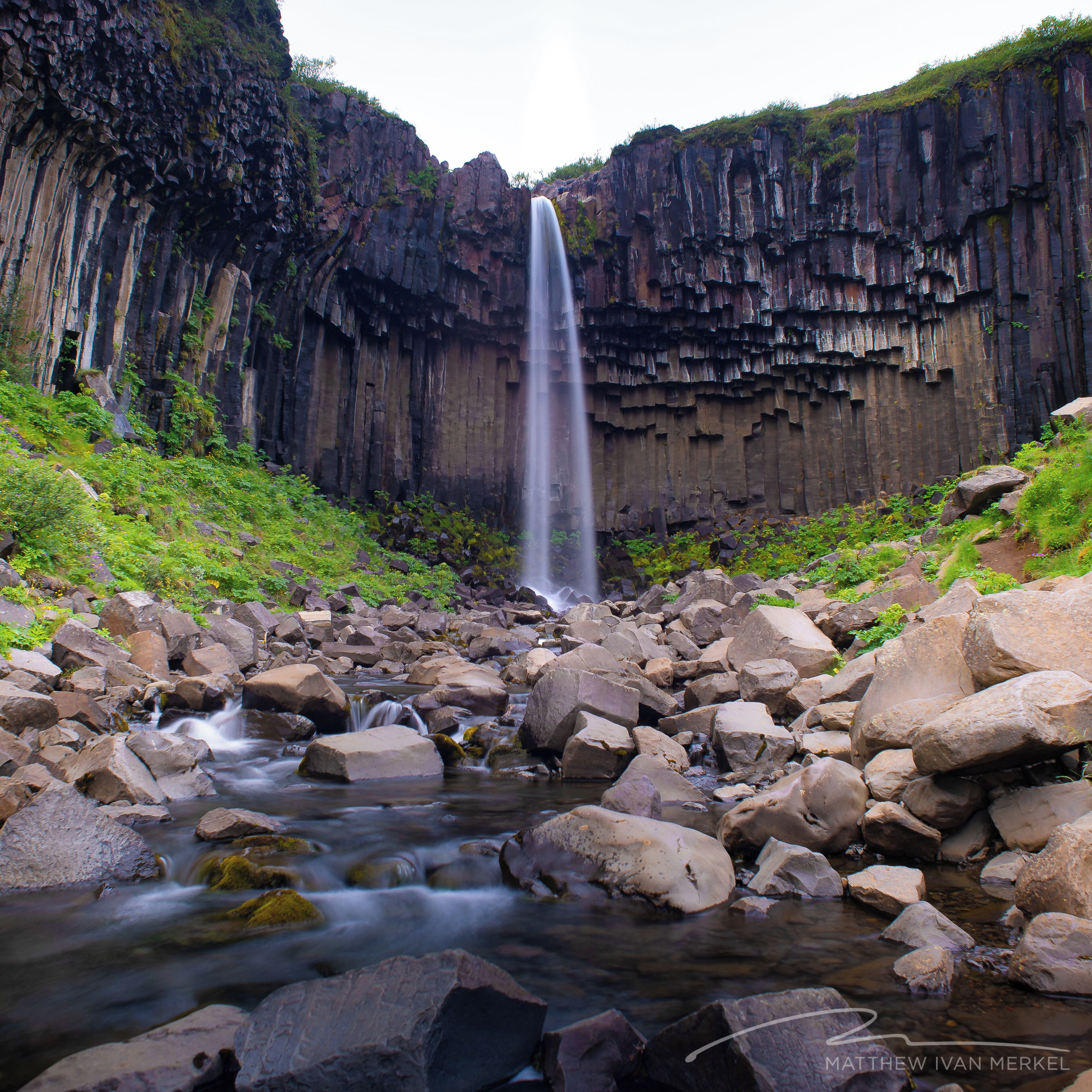 A waterfall cascading down a rocky cliff into a stream surrounded by boulders and greenery.