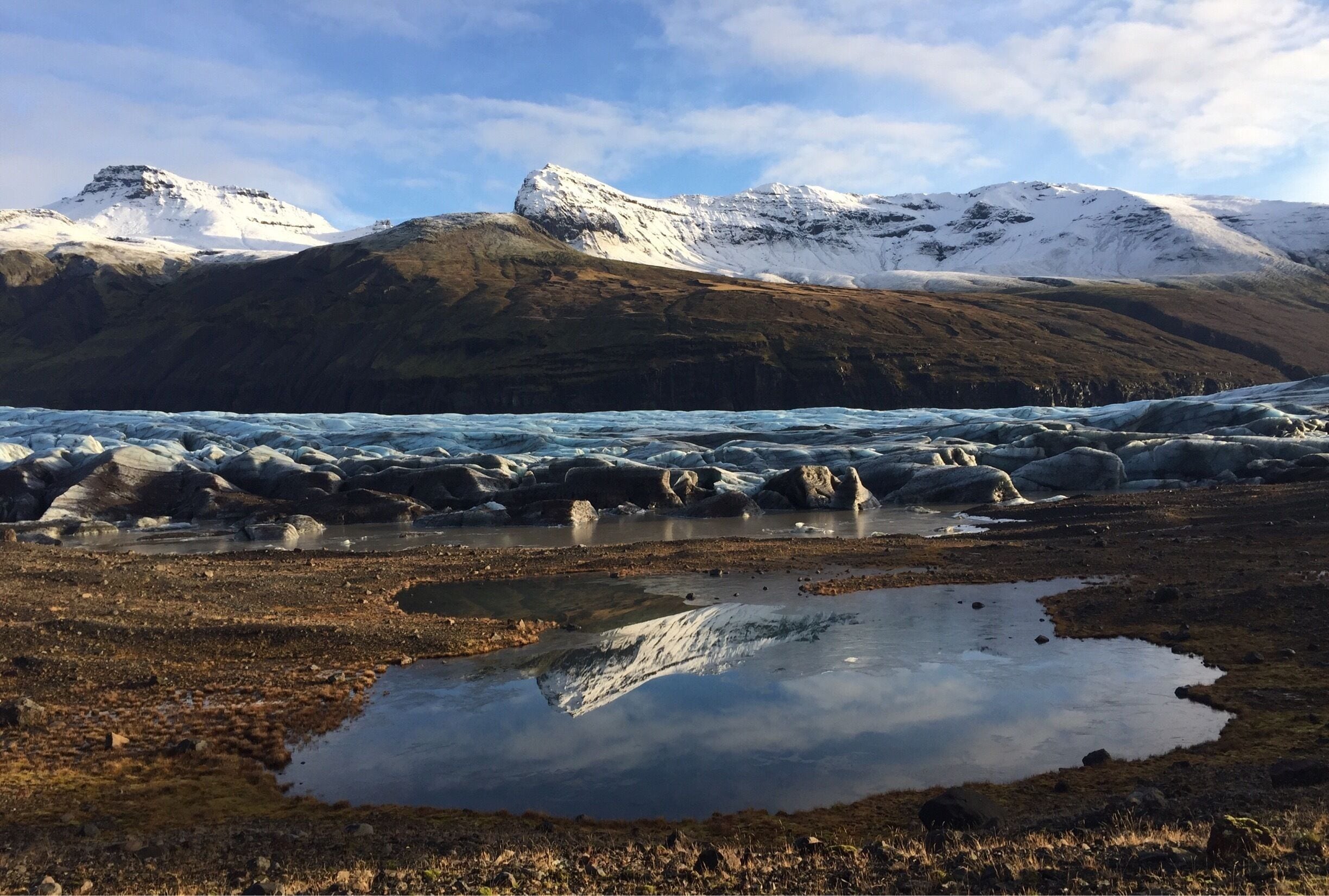 A glacier with a reflective pond, snow-capped mountains, and a rocky shoreline.