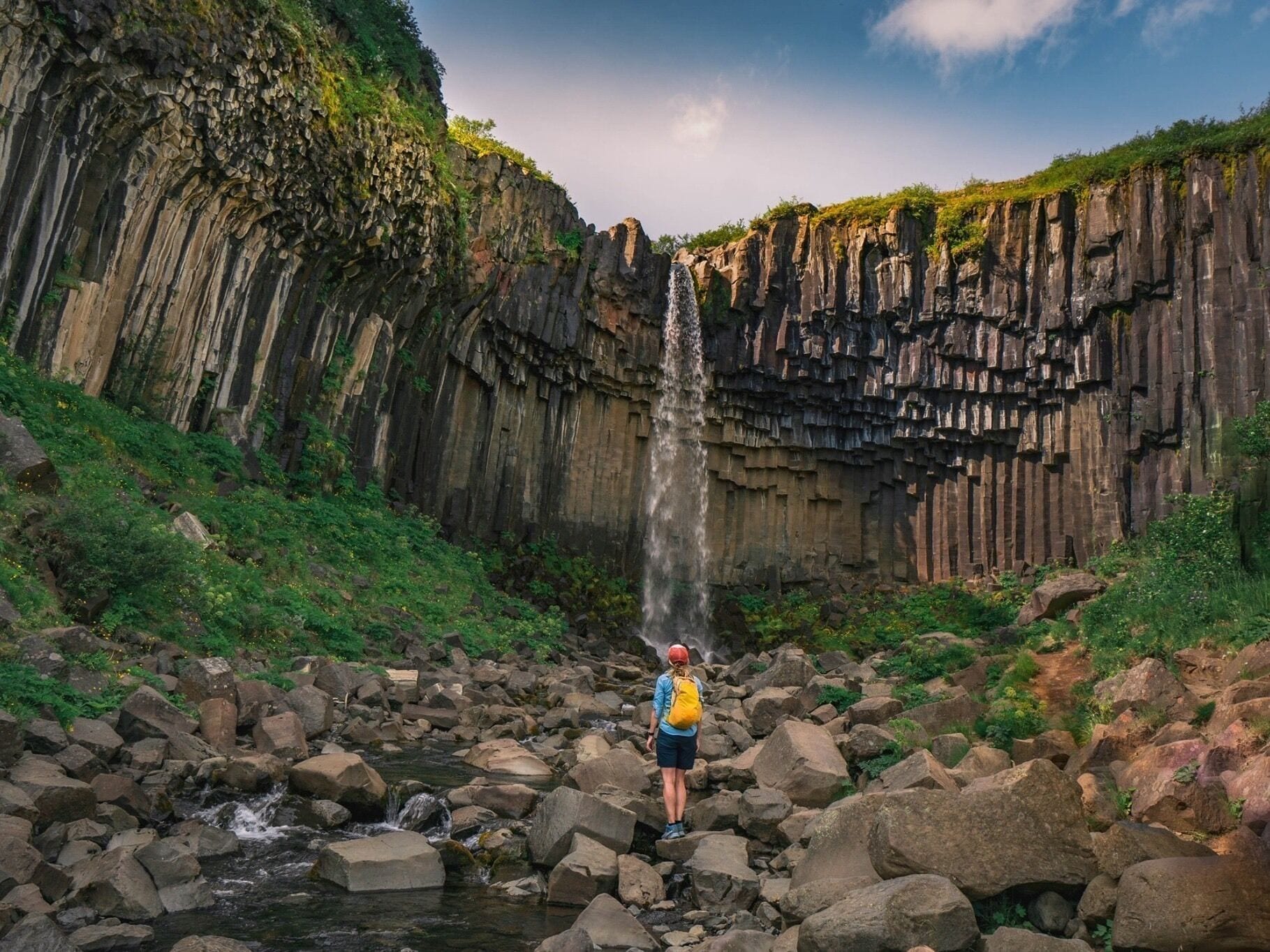 A waterfall cascading down a rocky cliff, surrounded by lush greenery and a hiker standing in the foreground.