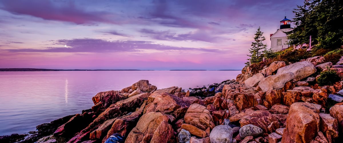 A rocky shoreline with a lighthouse and a calm sea under a colorful sky.