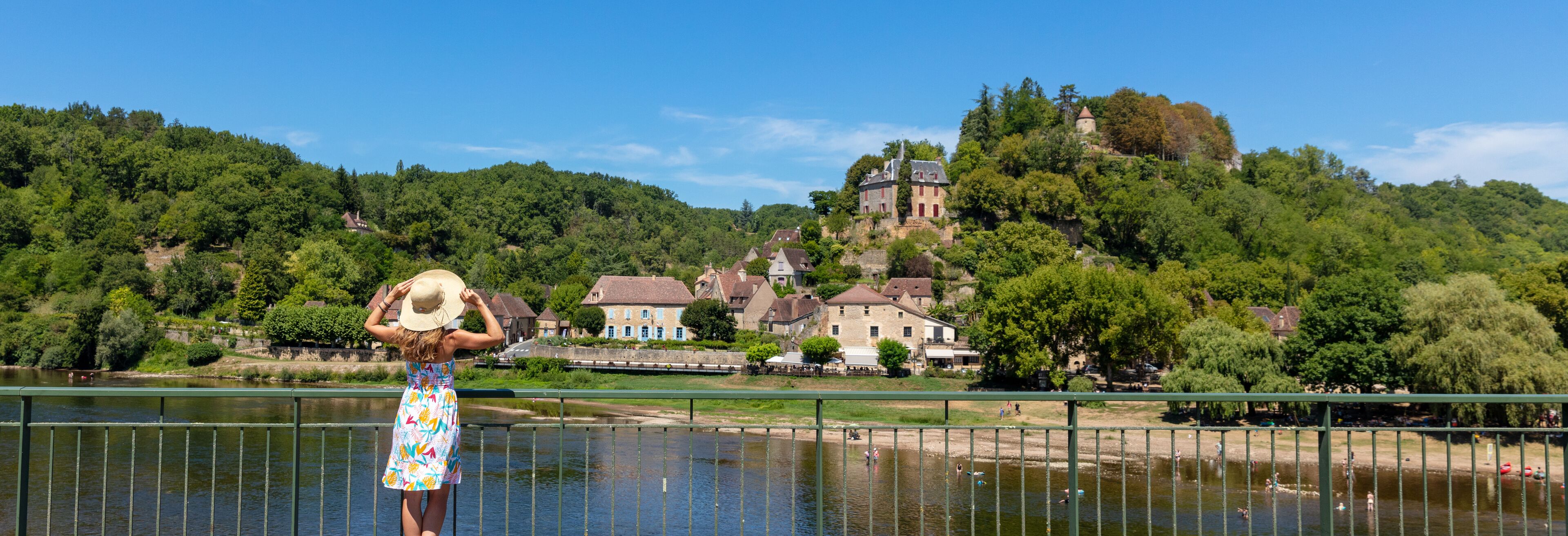 A woman in a colorful dress and straw hat standing by a river, overlooking a historic town with a castle on a hill.
