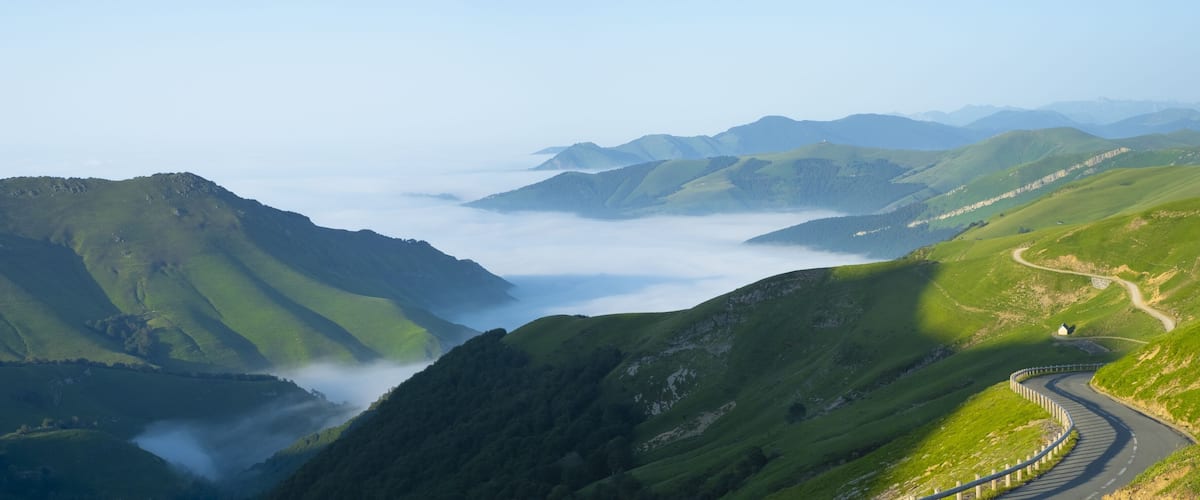 A winding mountain road surrounded by lush green hills and a valley shrouded in mist.
