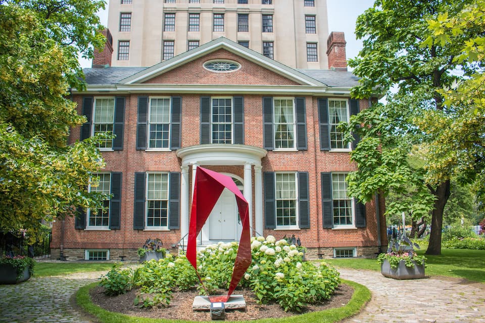 A red sculpture in front of a brick building with a white door and green shutters.
