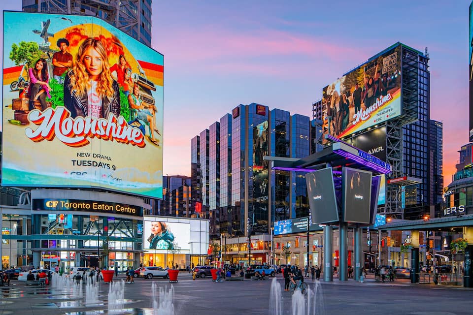 Uma praça urbana movimentada com um grande outdoor publicando 'Moonshine' e uma fonte no centro.